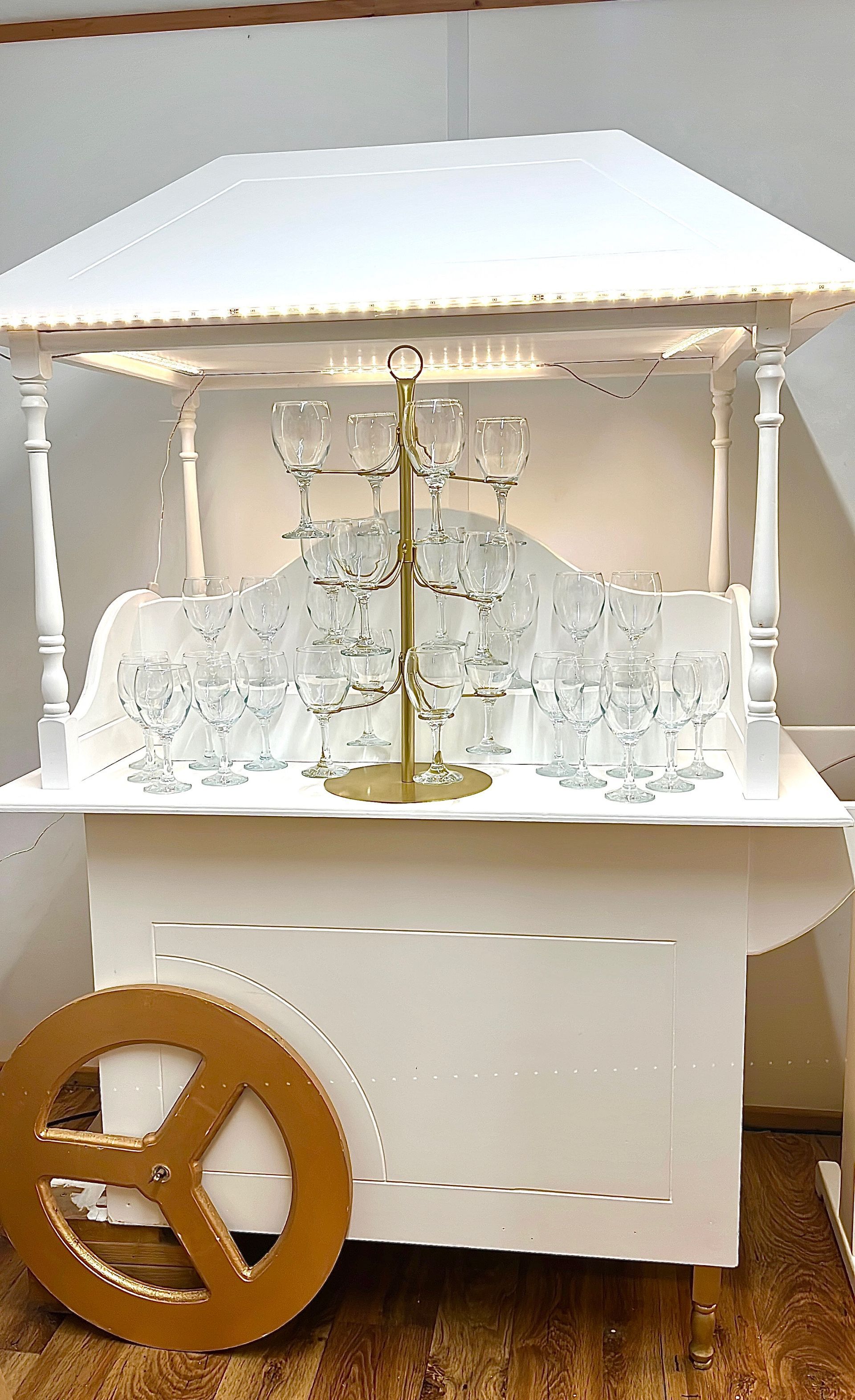 White miniature market stall with lace decor, hanging glasses, and a wooden wheel on the front