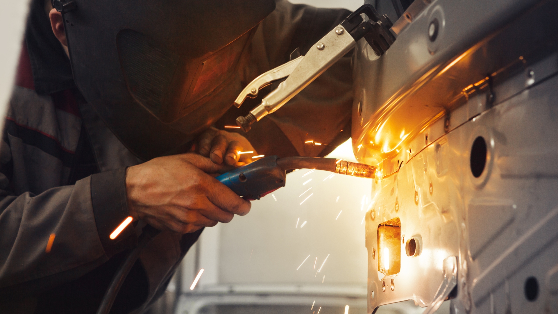 Worker welding a silver car body with a bright orange spark inside a garage