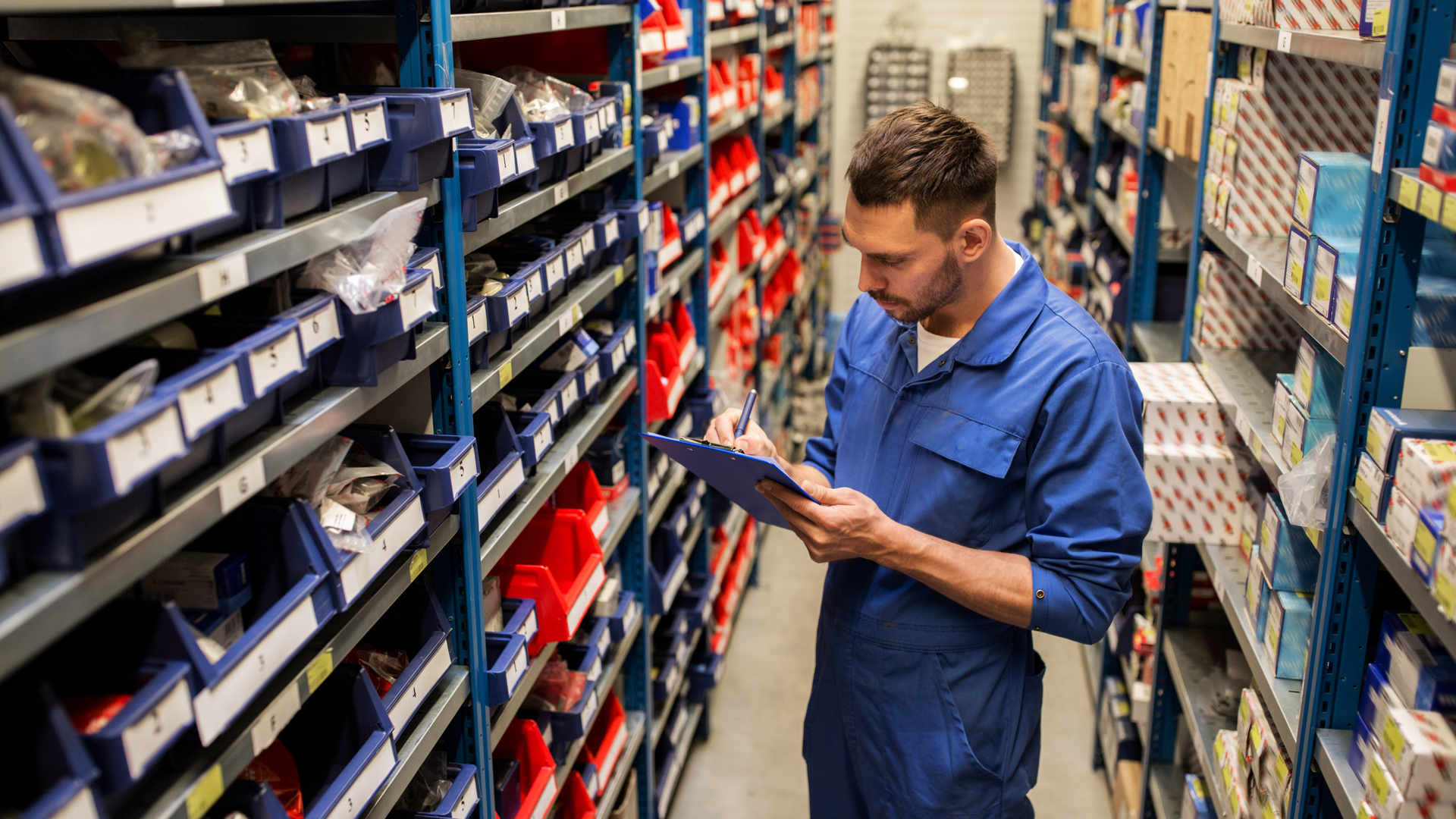 Worker in a blue uniform checking inventory in a warehouse aisle with shelves of labeled boxes