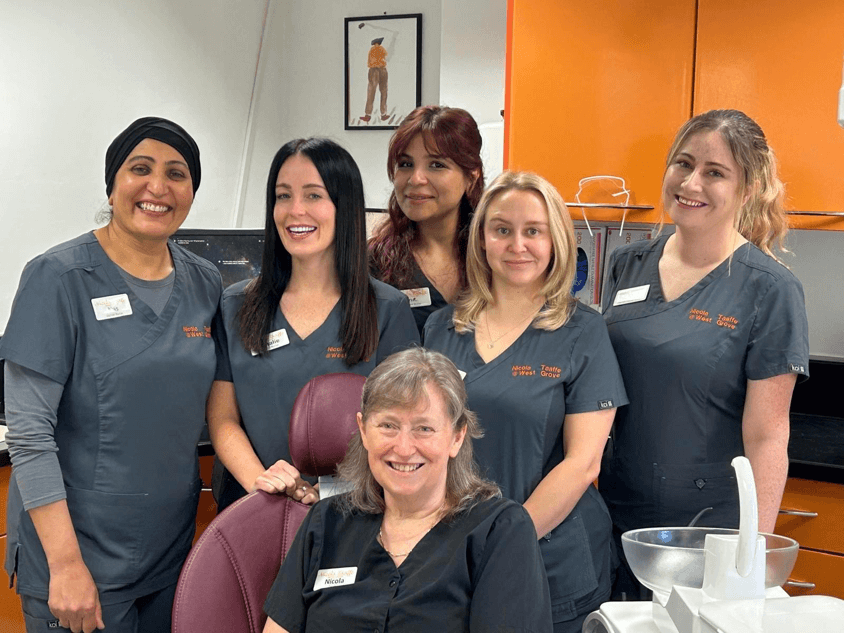 Six smiling staff members in gray uniforms pose in a bright clinic room with orange walls.