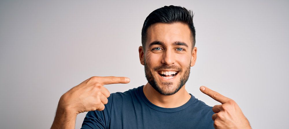 Smiling man in a dark shirt pointing at himself with both hands against a light gray background