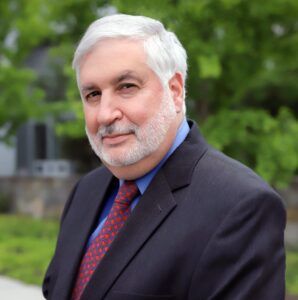 Man in a suit, with gray hair and beard, smiling outdoors in front of greenery.