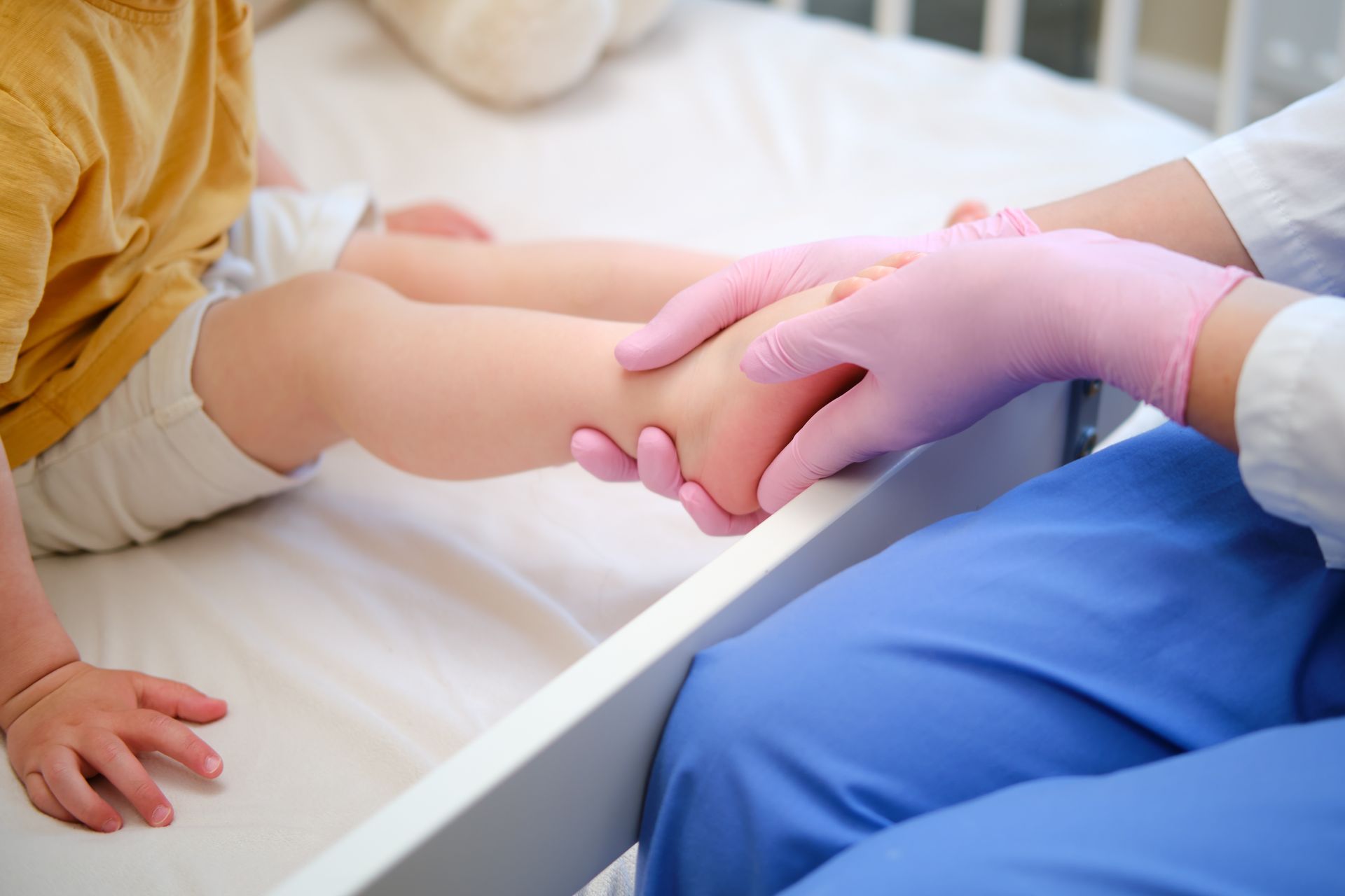 Healthcare worker examining a child's foot in bed, wearing gloves and blue scrubs.