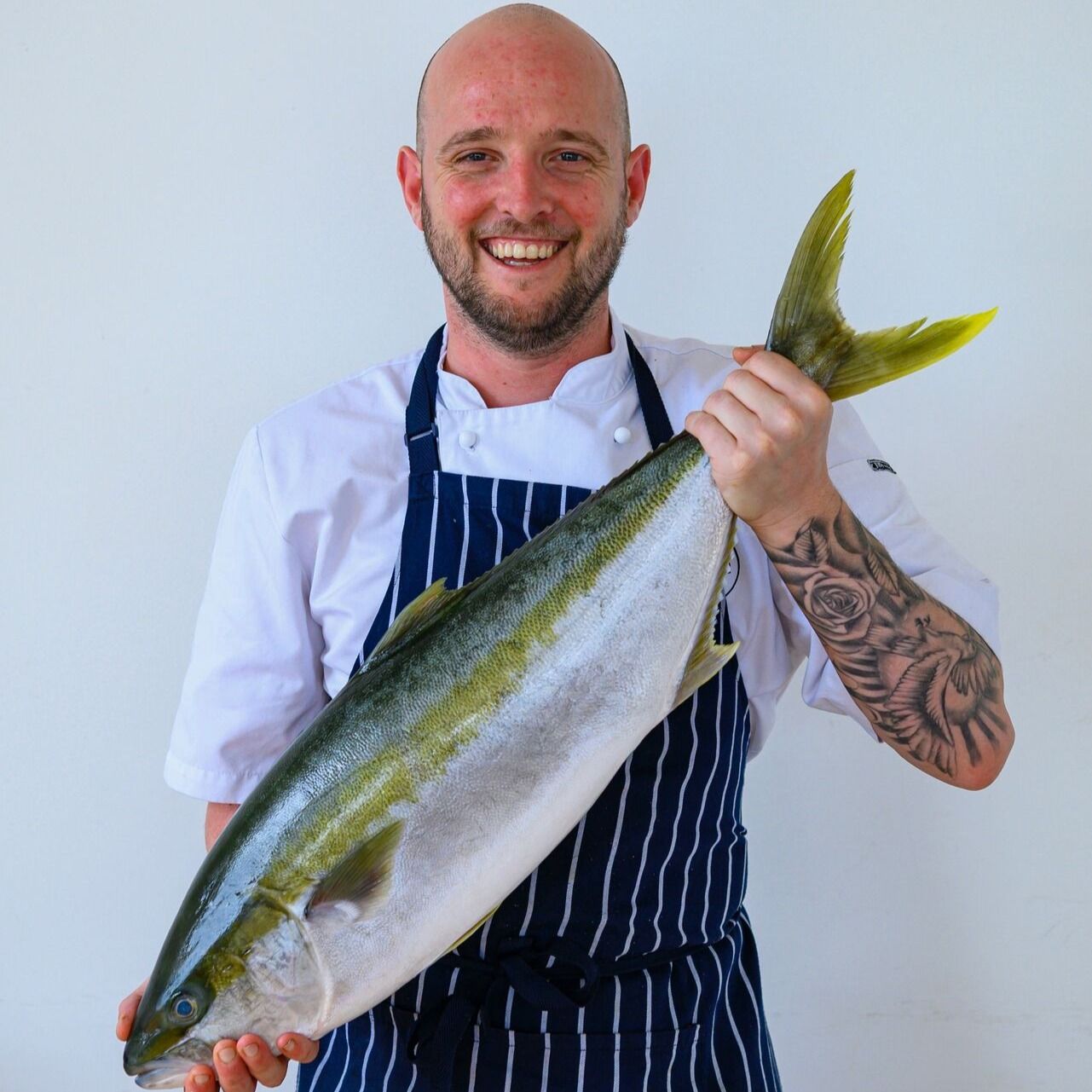 Chef holding a large yellowtail fish, smiling against a plain white background.