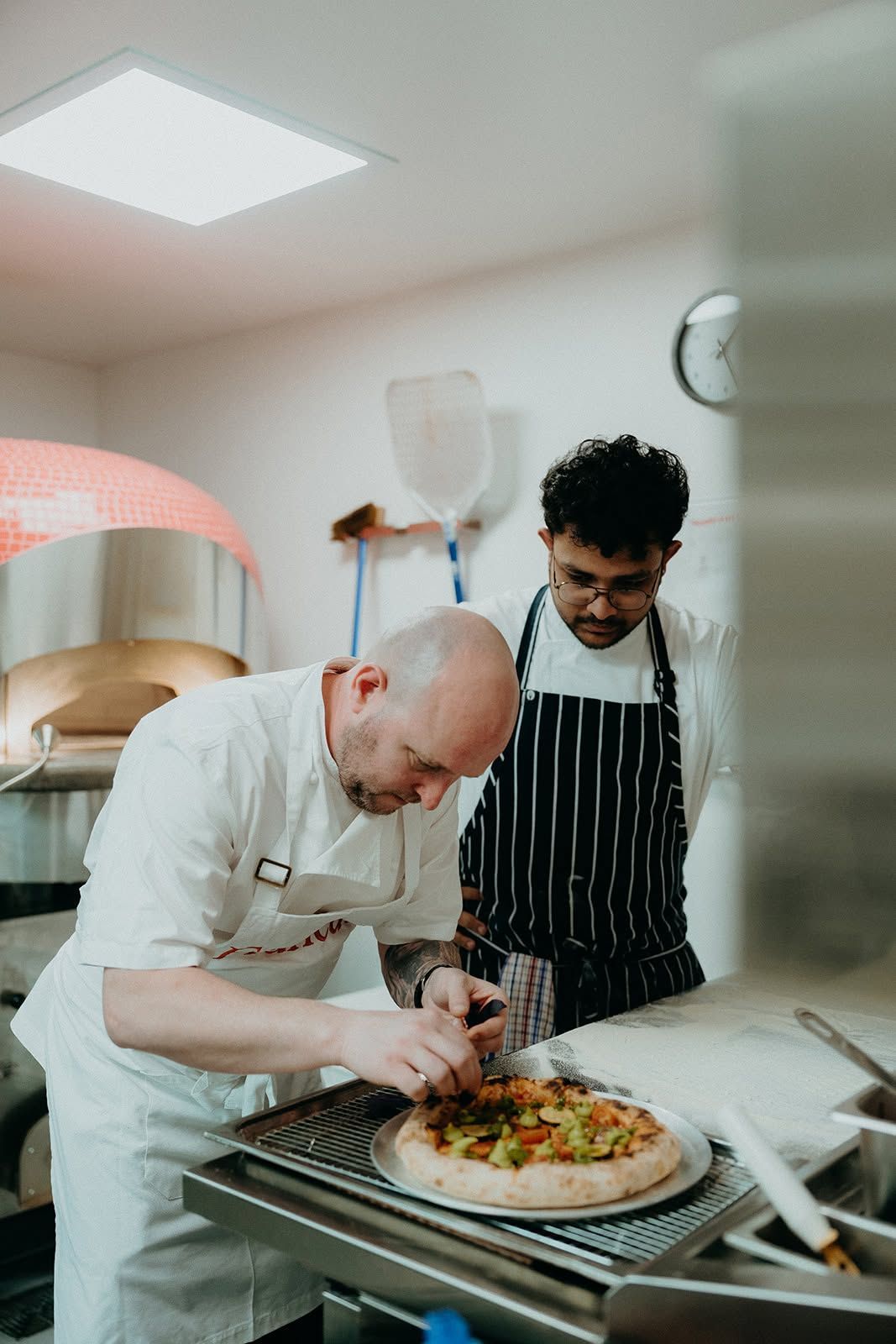Two chefs in a kitchen plating a pizza together, one in white and one in a striped apron.