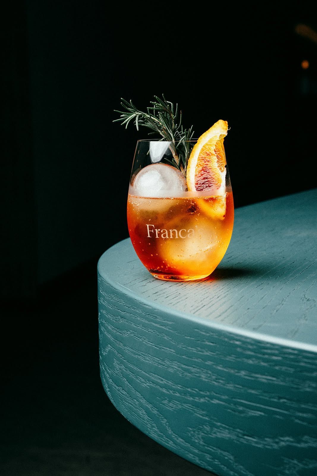 Orange cocktail with ice, grapefruit slice, and garnish on a teal round table against a dark background