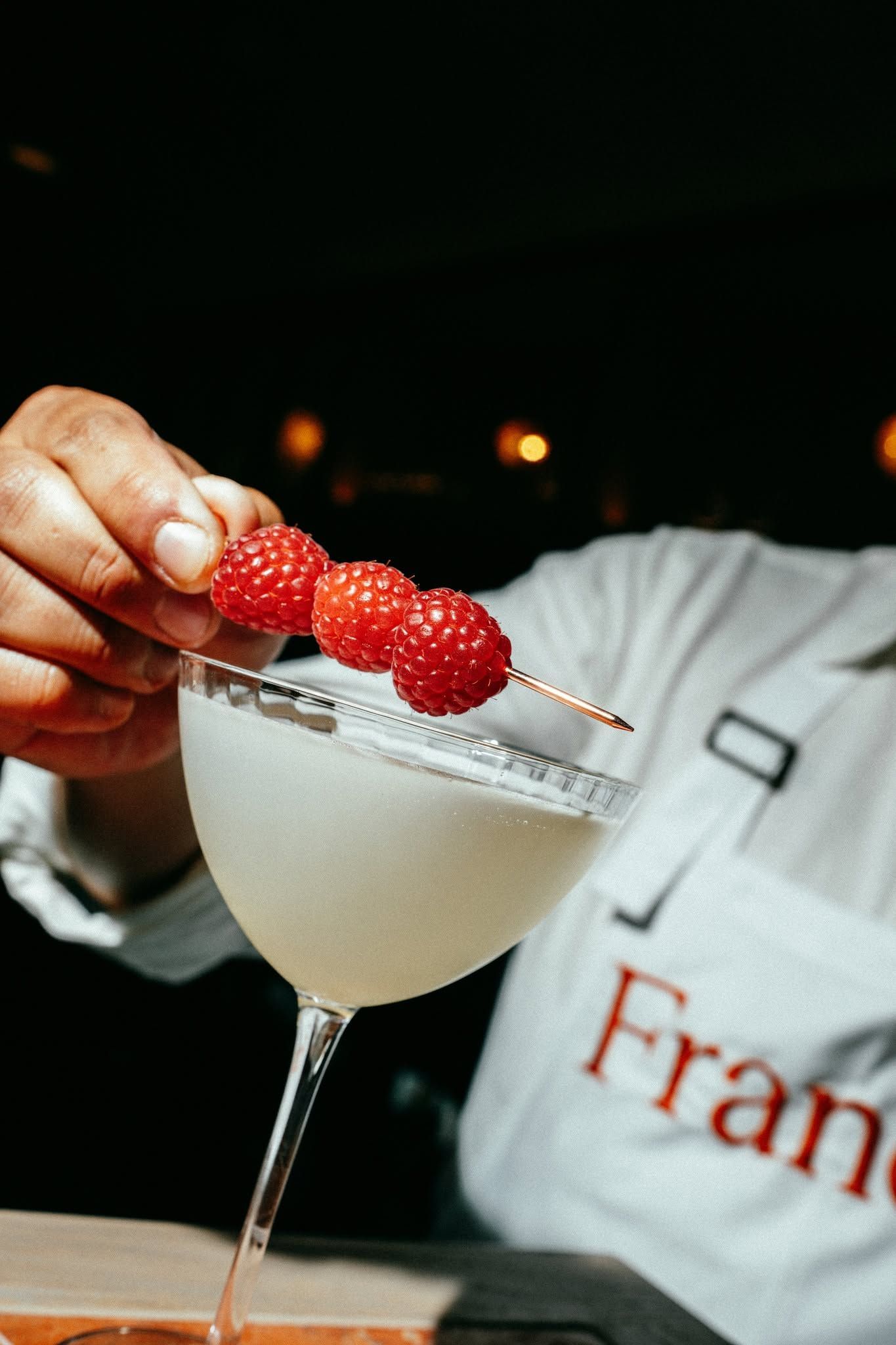Hand placing raspberries on a sugared cocktail glass in a dim bar setting
