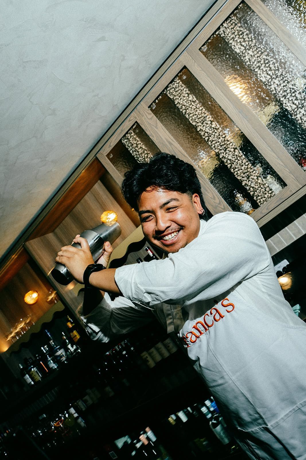 Smiling person in a white shirt holding a bottle in a dim bar with shelves of drinks behind them