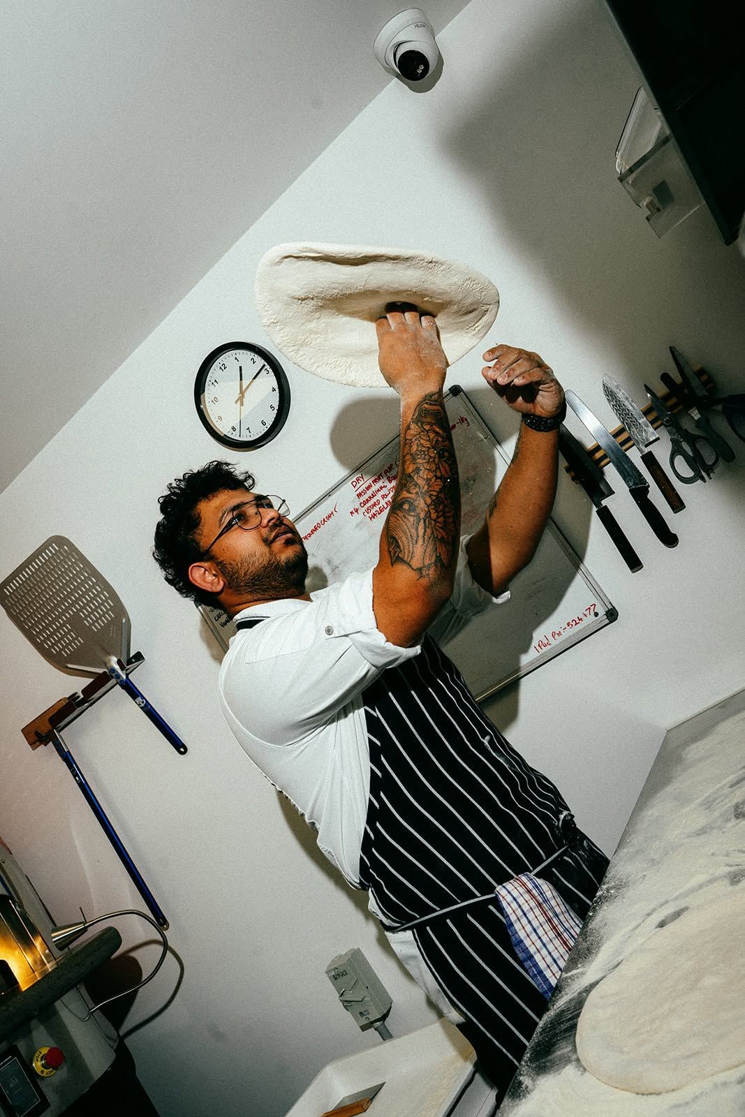 Chef tossing dough in a kitchen with a wall clock and hanging utensils