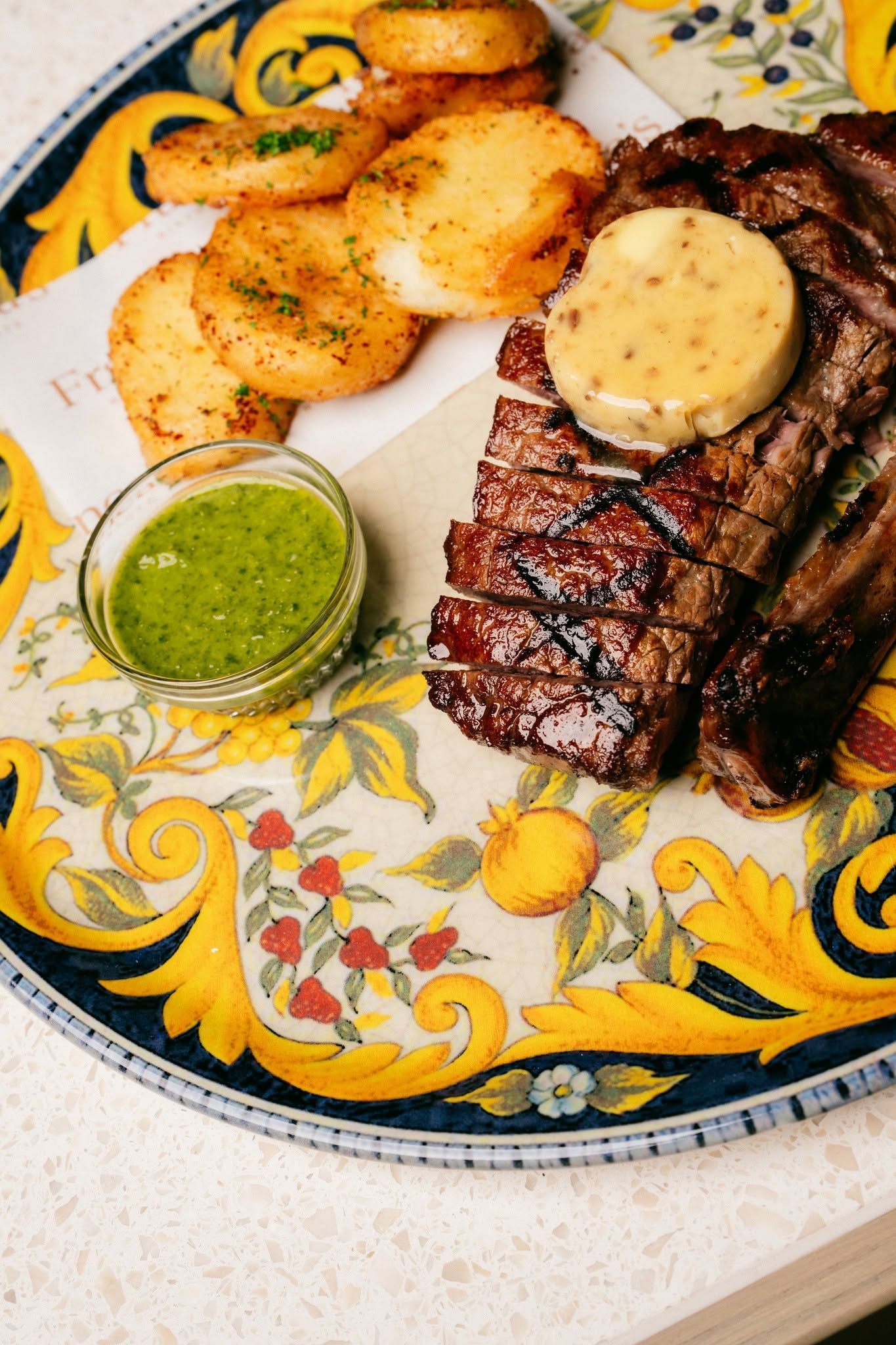 Grilled steak with herb sauce, roasted potatoes, and green chutney on a patterned plate