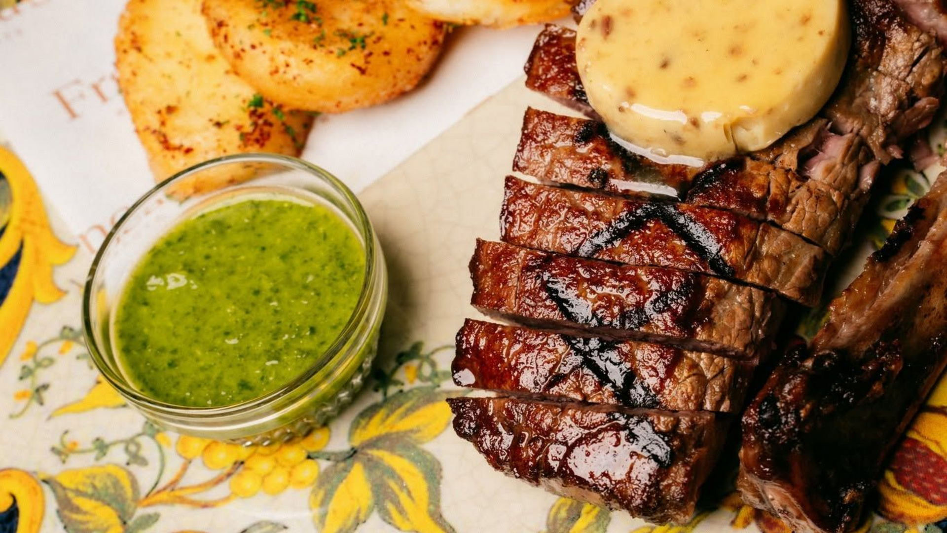Grilled steak with sauce, potato wedges, and green chutney on a floral plate