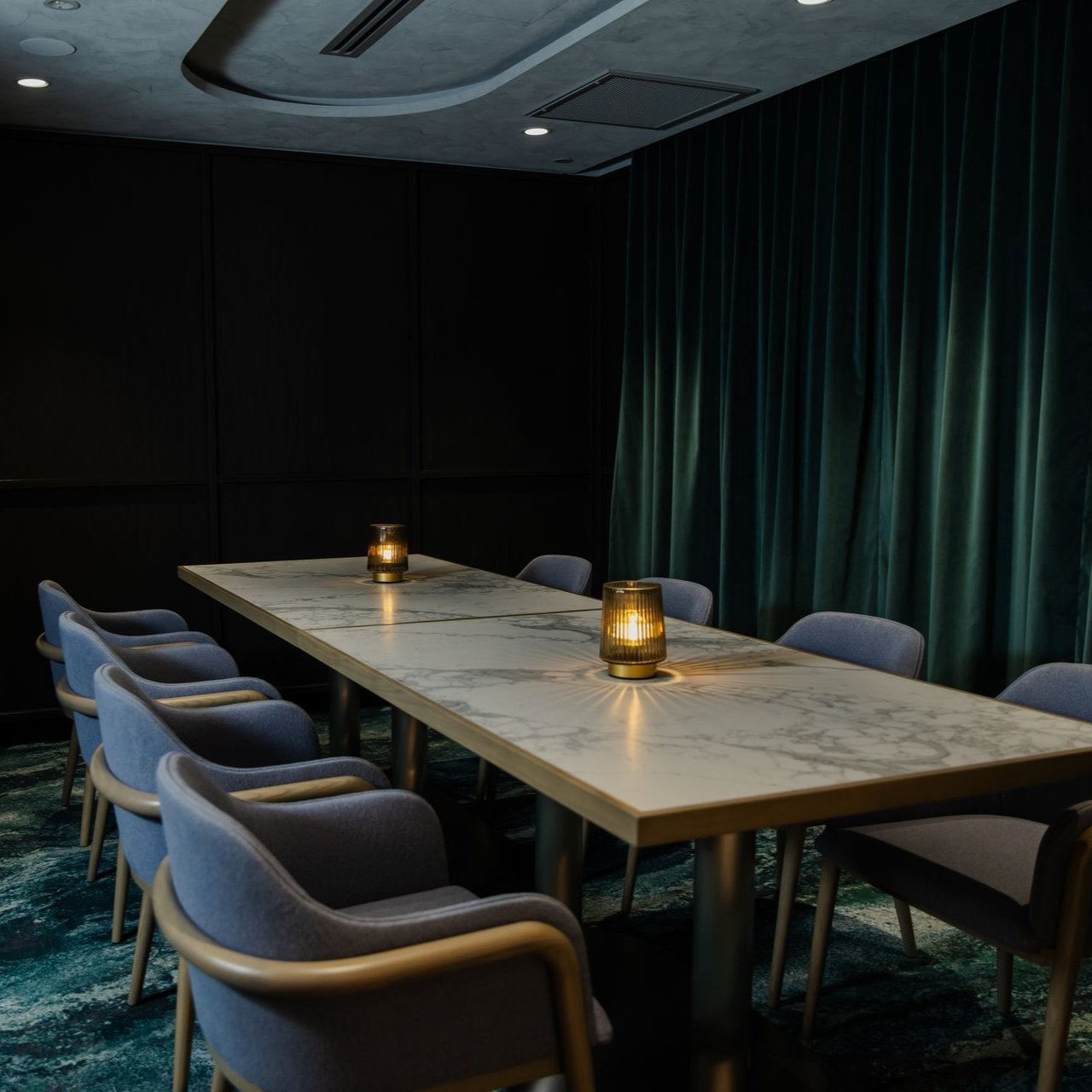 Private dining room with a long wooden table, blue chairs, and warm candle-like lights by dark teal curtains