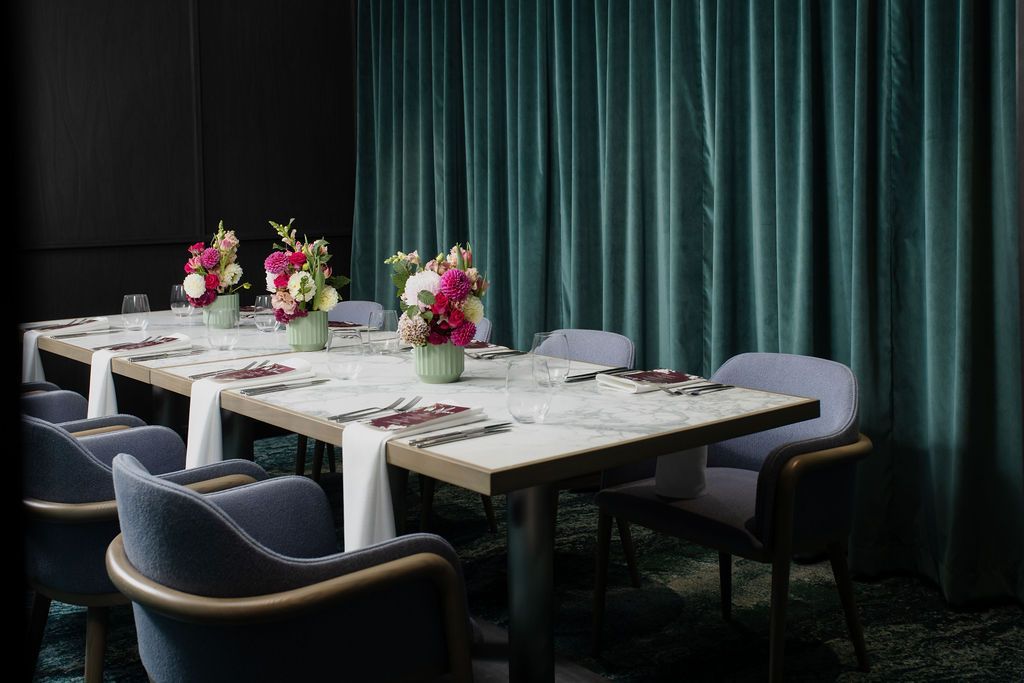 Private dining room with a long wooden table, blue chairs, and warm candle-like lights by dark teal curtains