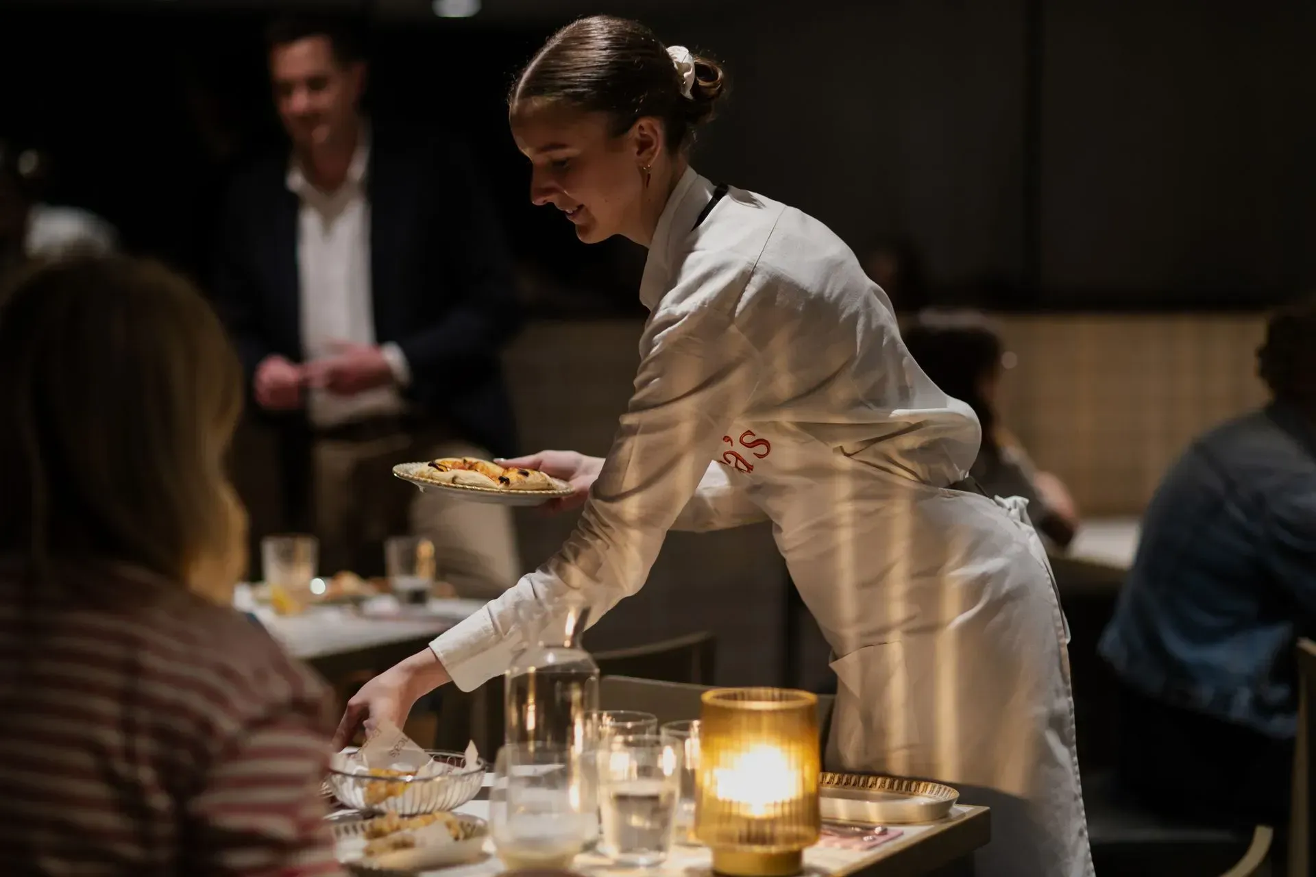 Server in a white uniform serving dessert at a dimly lit restaurant table with candles