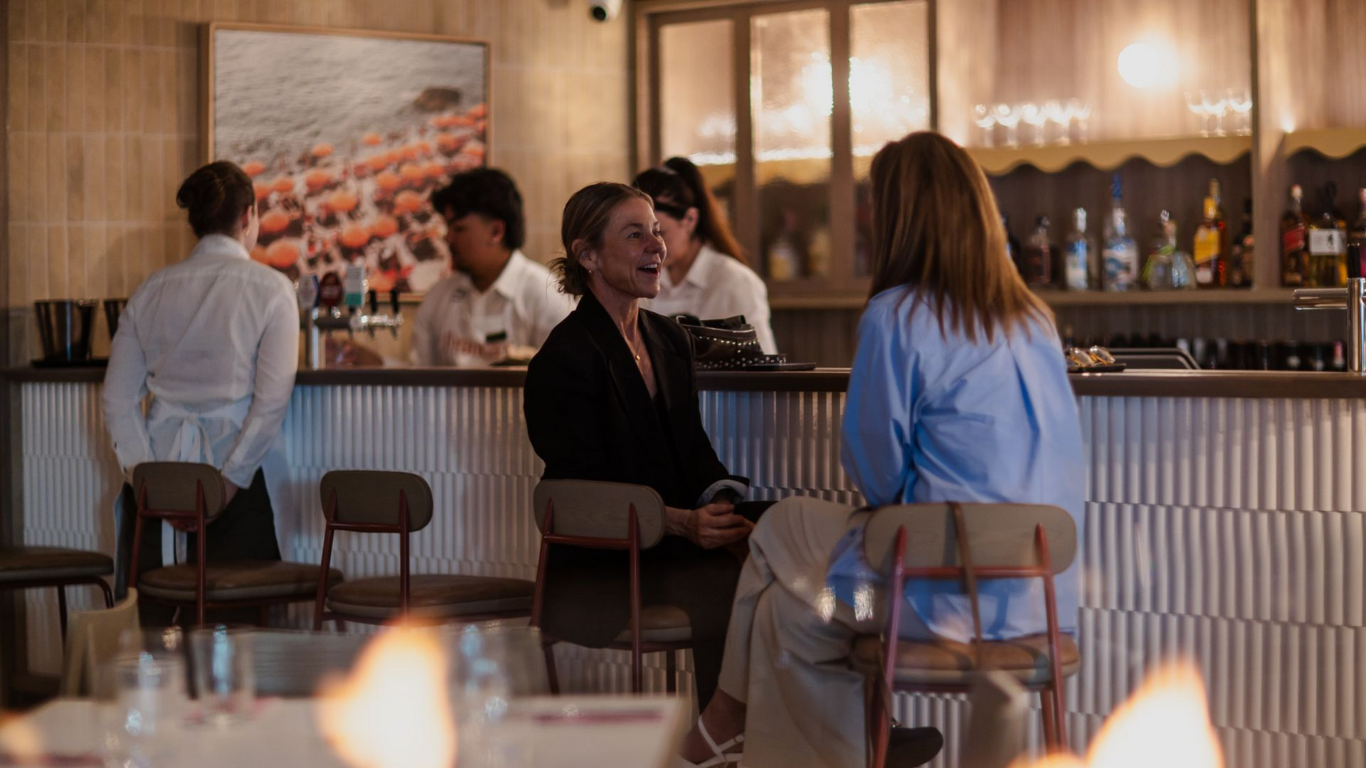 People chatting at a warmly lit bar with bottles, stools, and a floral wall artwork in the background.