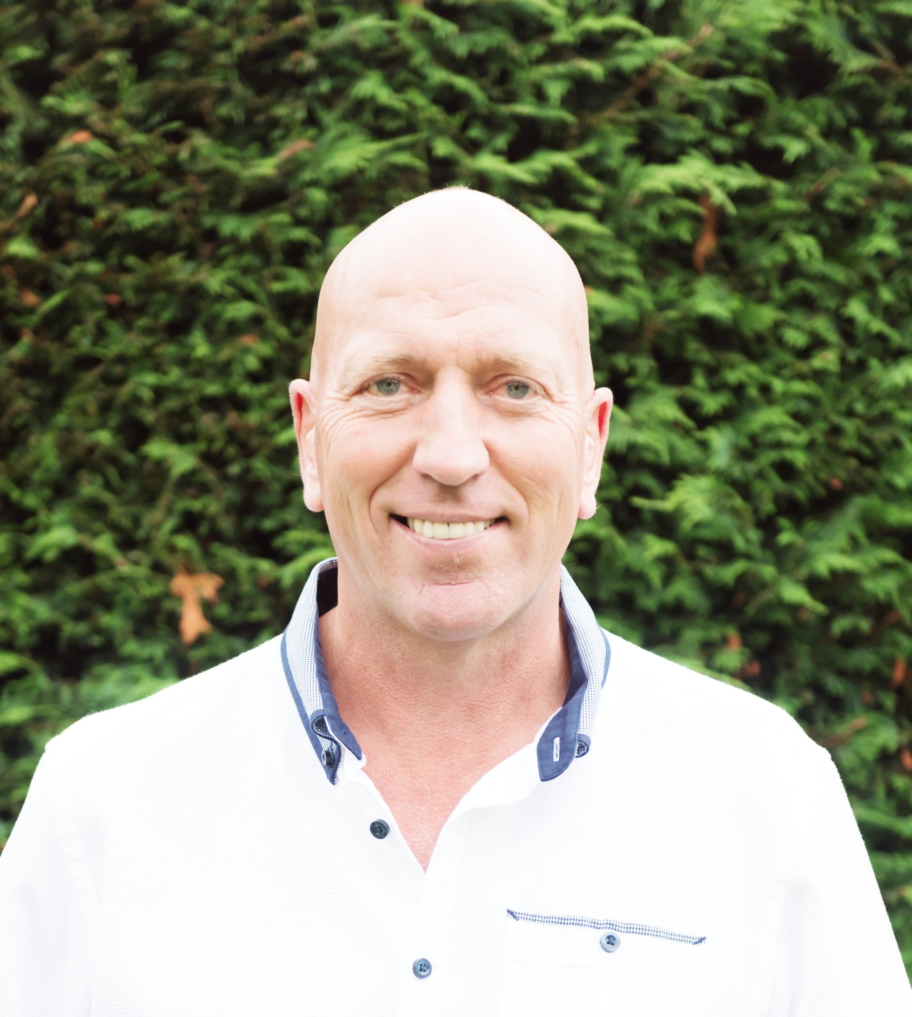 Roy in a white shirt is smiling in front of a bush.
