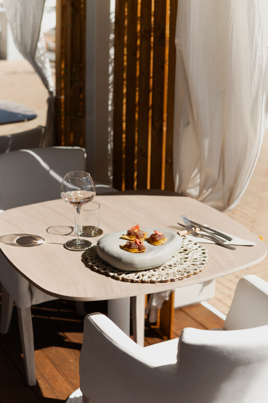 Elegante tavolino da caffè con dessert su un piatto decorato accanto a un calice di vino e sedie bianche vicino a una finestra.