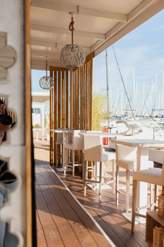 Caffè luminoso sul porto turistico con tavoli e sedie bianche accanto alle barche a vela ormeggiate.