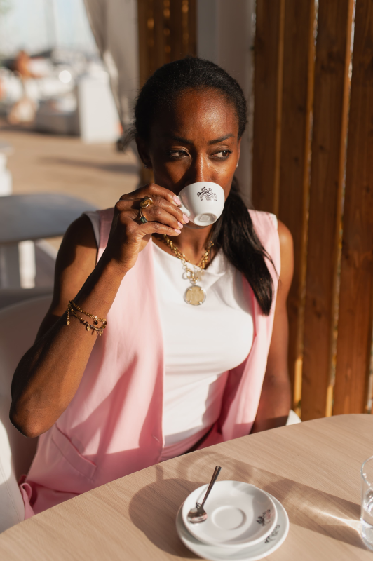 Una donna con un gilet rosa sorseggia un caffè a un tavolino di un bar illuminato dal sole.