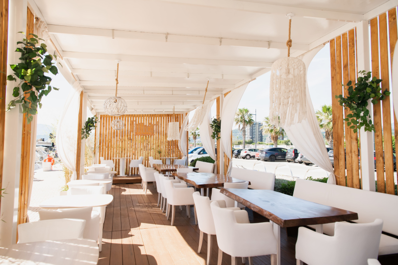 Luminoso patio esterno con tavoli in legno, sedie bianche e piante sospese, immerso nella luce del sole.