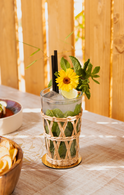 Un bicchiere di vetro intrecciato contenente una bevanda ghiacciata alla menta, un girasole e una cannuccia nera, su un tavolo di legno illuminato dal sole.