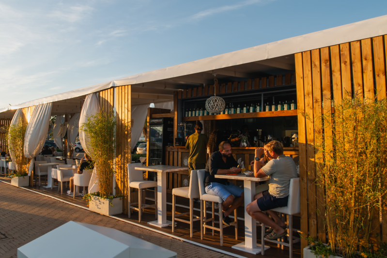 Bar e lounge all'aperto al tramonto, con persone sedute al bancone sotto una moderna tettoia bianca e pareti in legno.