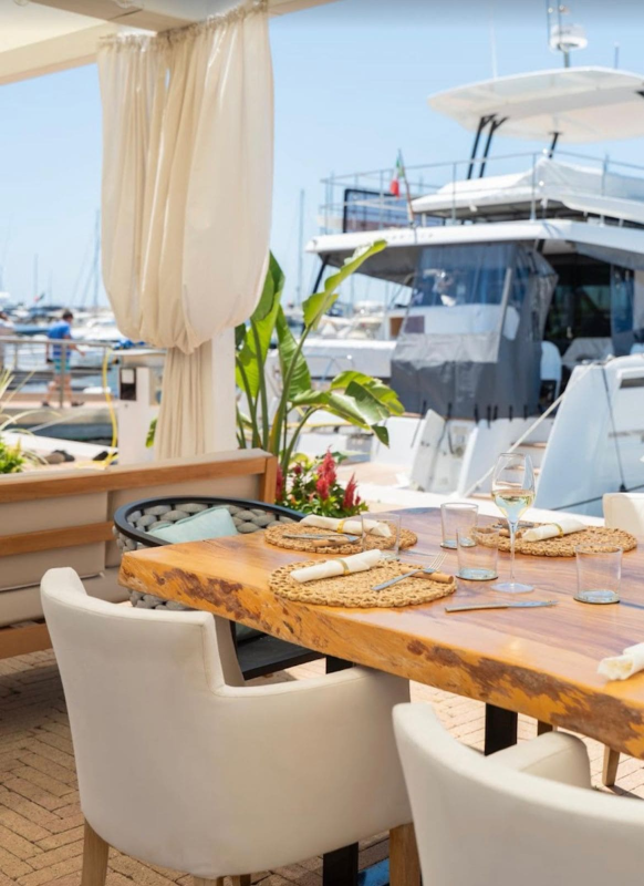 Tavolo da pranzo su uno yacht, su un ponte ombreggiato, con tovagliette intrecciate, sedie bianche e vista sul porto turistico.