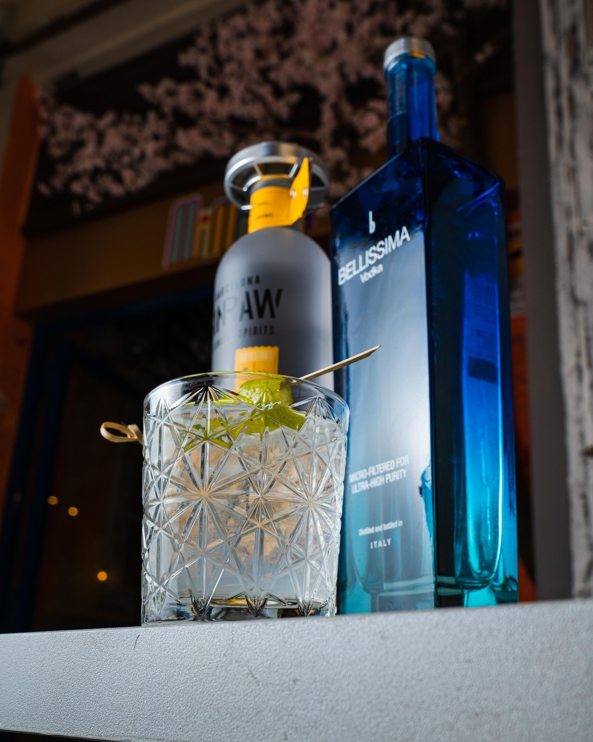 Un bicchiere di cristallo con ghiaccio e limone accanto a due bottiglie di liquore blu sul bancone del bar.