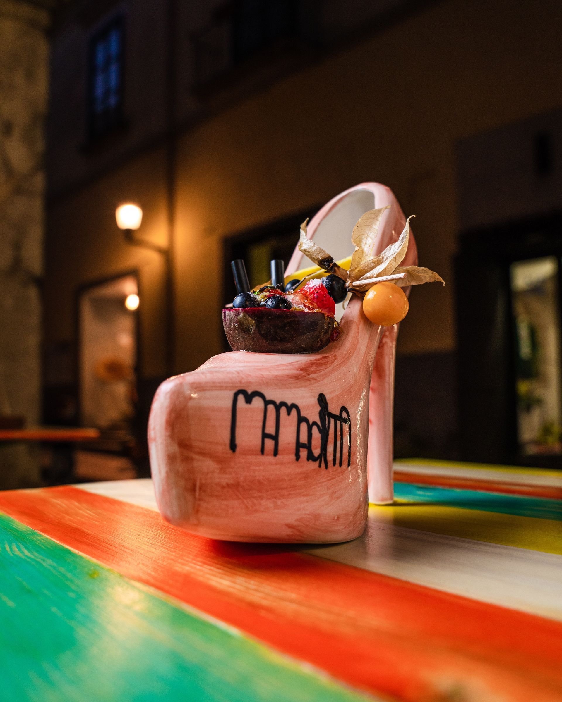 Cocktail rosa in un bicchiere su un tavolo colorato, guarnito con frutta, in un'atmosfera da bar con luci soffuse.
