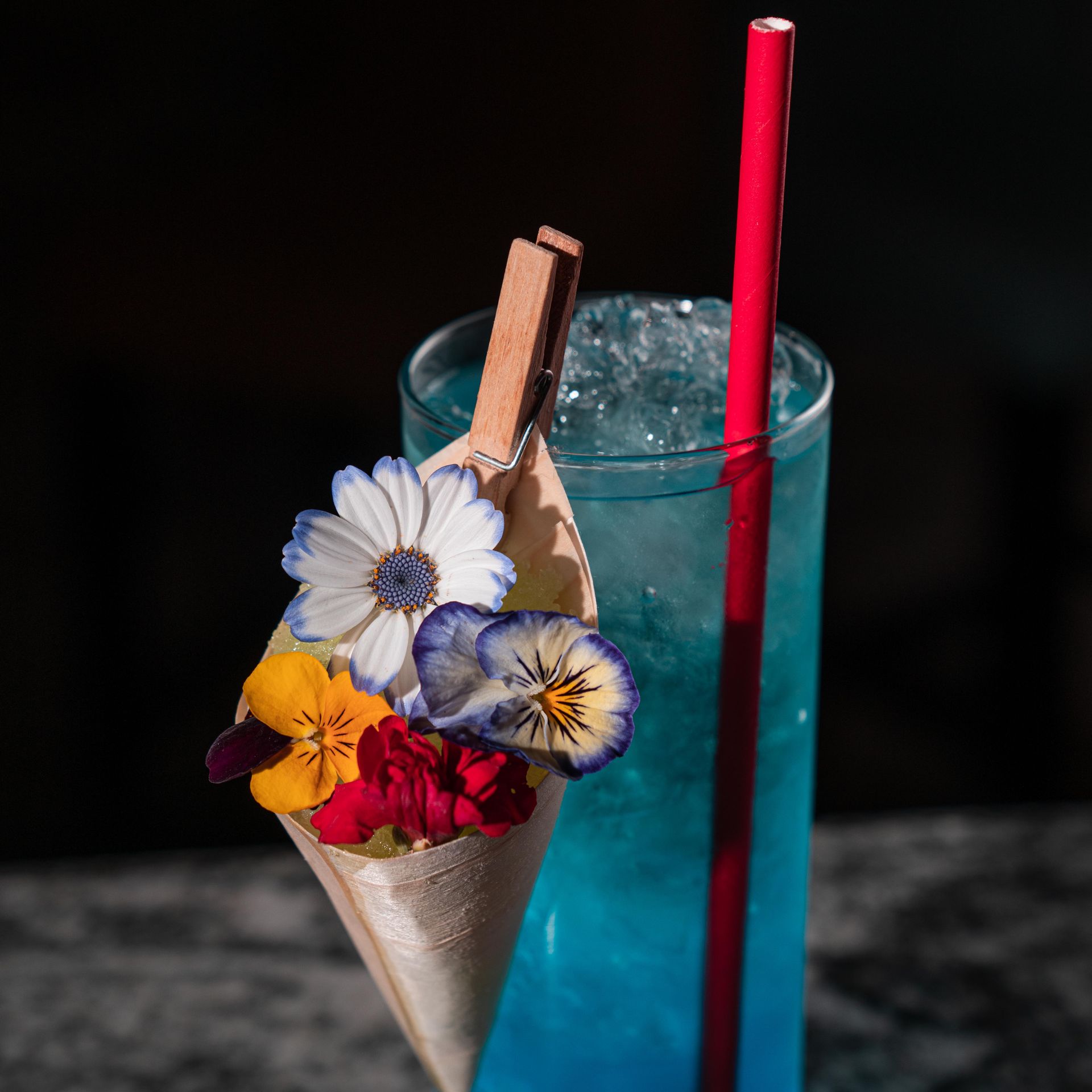 Cocktail blu in un bicchiere a cono con fiori e bastoncini di cannella accanto a una cannuccia rossa.