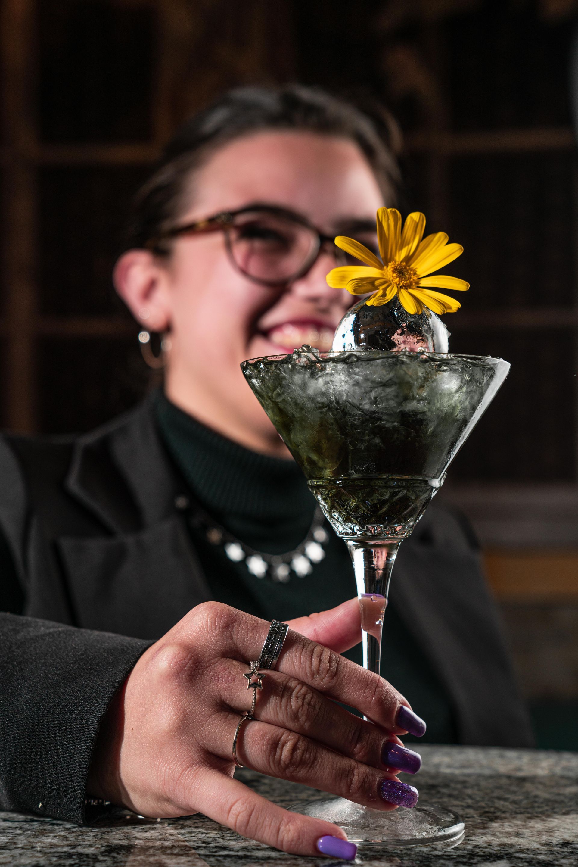 Una persona, in un bar in penombra, tiene in mano un bicchiere da martini guarnito con un fiore giallo e sorride.