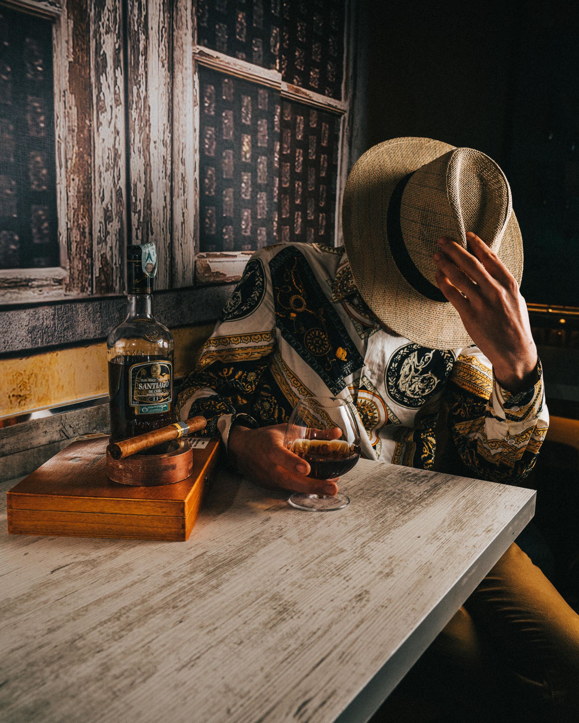 In una stanza in penombra, una persona con indosso una vestaglia a motivi e un cappello di paglia siede a un tavolo con una bottiglia di whisky.