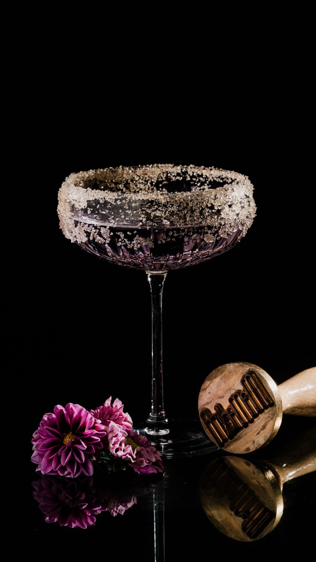 Cocktail scuro in una coppa con un fiore rosa e uno shaker in legno su sfondo nero.