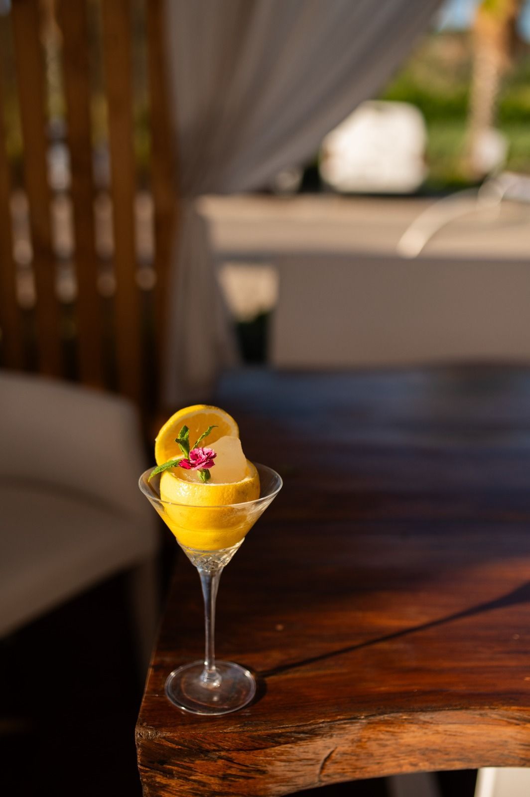 Cocktail giallo in un bicchiere con stelo e guarnizione floreale su un tavolo di legno all'aperto.