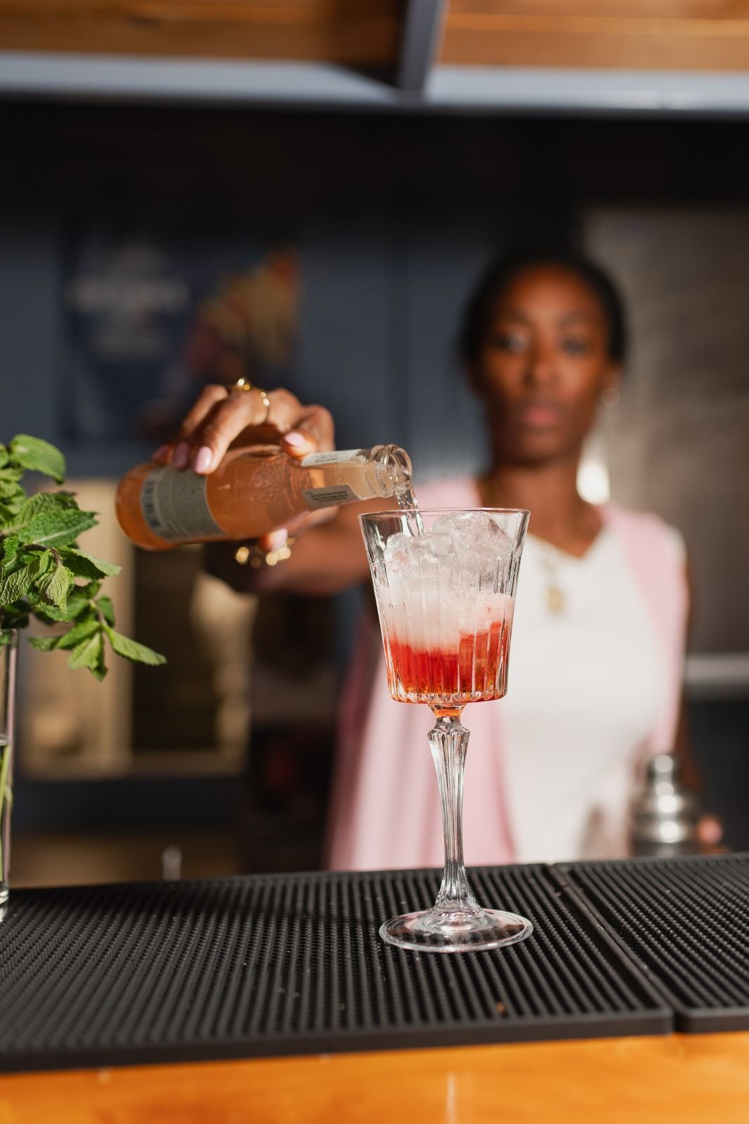 Al bancone di un bar, un barista versa un drink in un bicchiere alto, con uno shaker sfocato sullo sfondo.