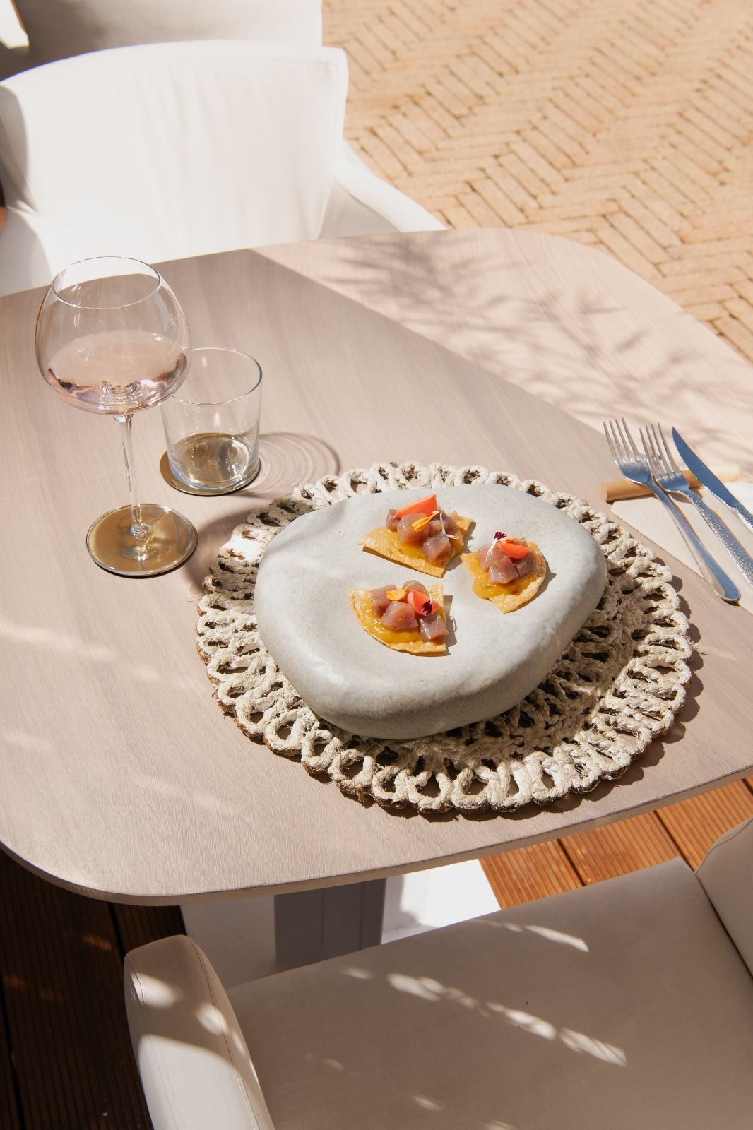Tavolo all'aperto con piatto da dessert, bicchiere da vino e bibita piccola, esposto al sole su un patio.
