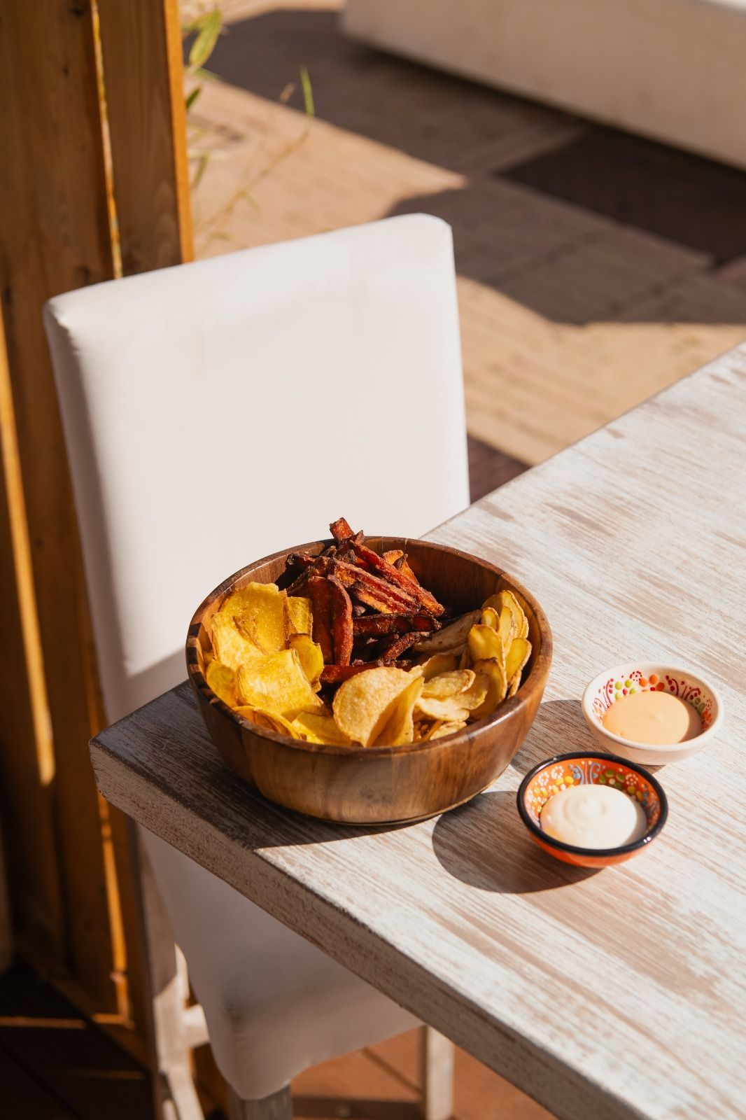 Una ciotola di patatine fritte con sopra cibi fritti e guarnizione rossa, servita su un tavolo all'aperto illuminato dal sole, con salse per intingere.