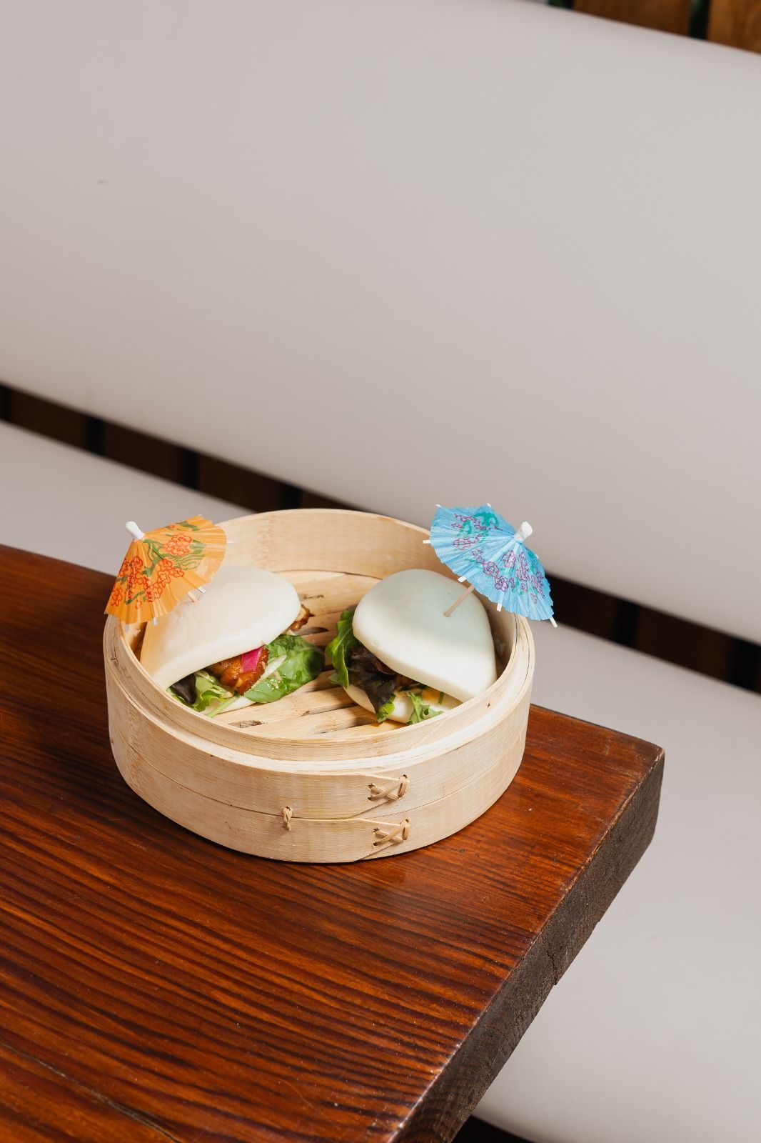 Un piccolo cestino di bambù con dim sum e ombrellini di carta colorati su un tavolo di legno.