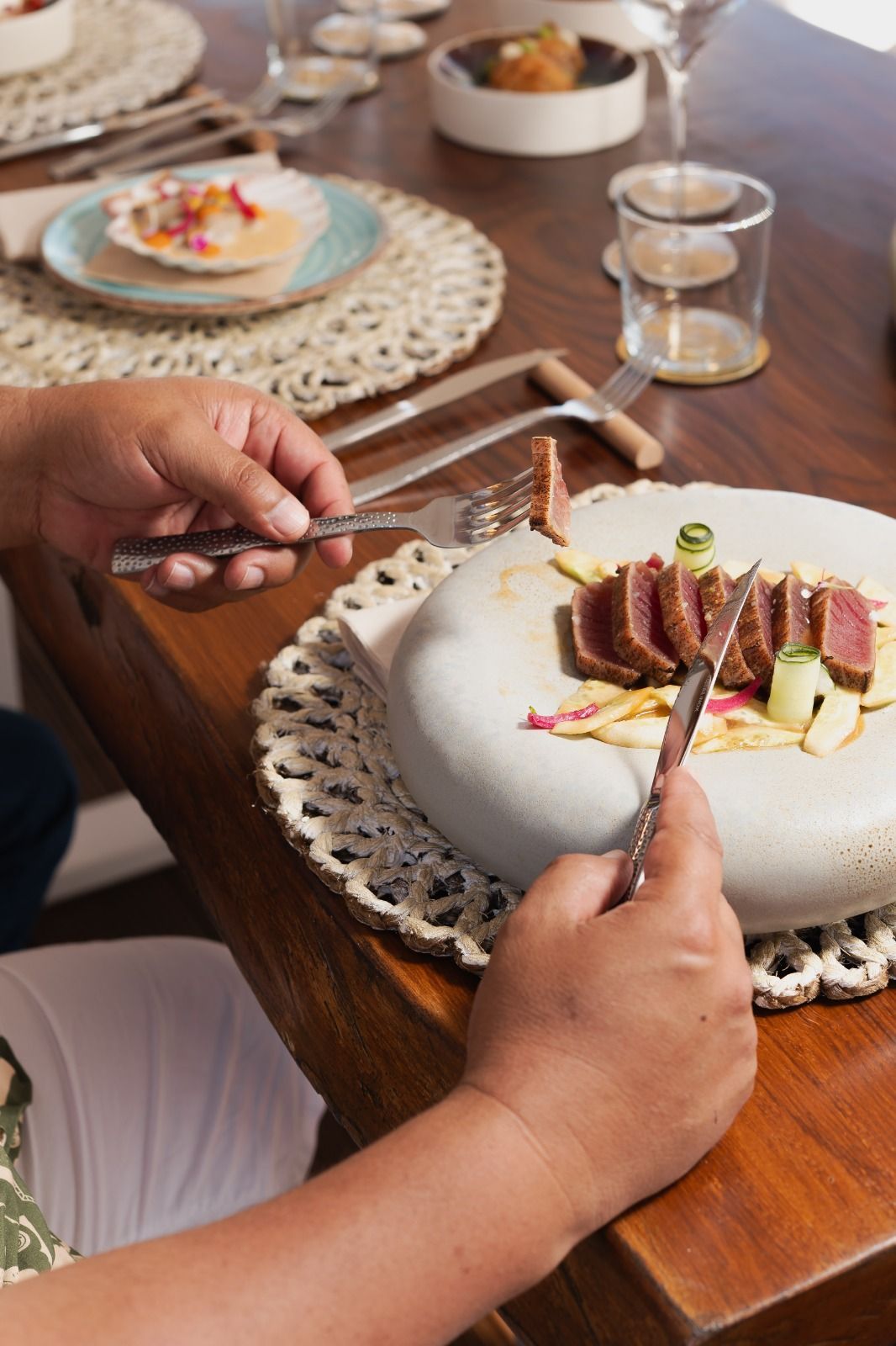 Mani che servono una bistecca su un piatto bianco a un tavolo di legno con tovagliette intrecciate e bicchierini.