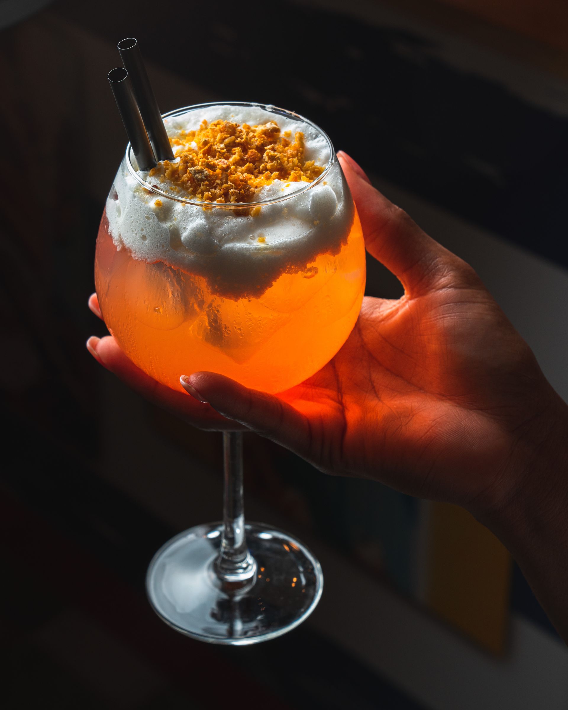 Mano che regge un bicchiere da cocktail con stelo, contenente una bevanda arancione brillante, ghiaccio e una spuma zuccherata in superficie.
