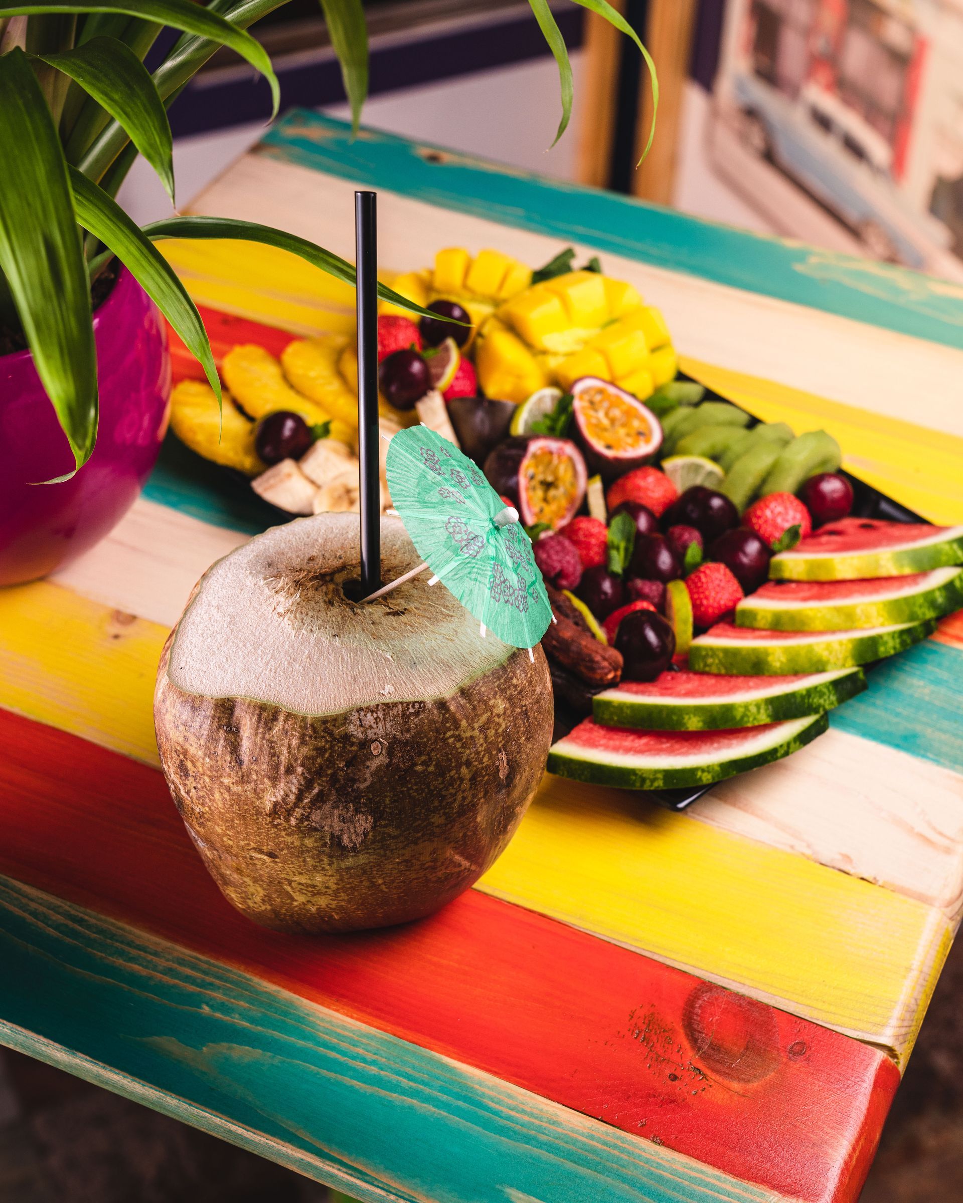 Piatto di frutta tropicale con anguria, frutti di bosco, banane e una bevanda al cocco su un tavolo a righe.