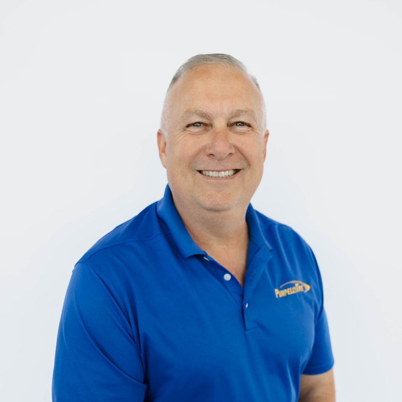 A smiling man wearing a bright blue polo shirt with a gold logo, positioned against a plain, off-white background.