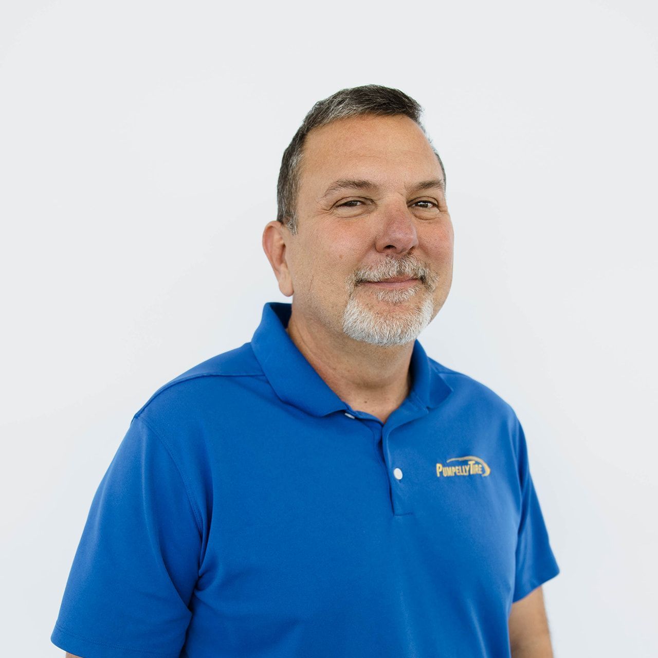 A person with a goatee wearing a bright blue polo shirt with a gold logo on the chest, standing against a white background.