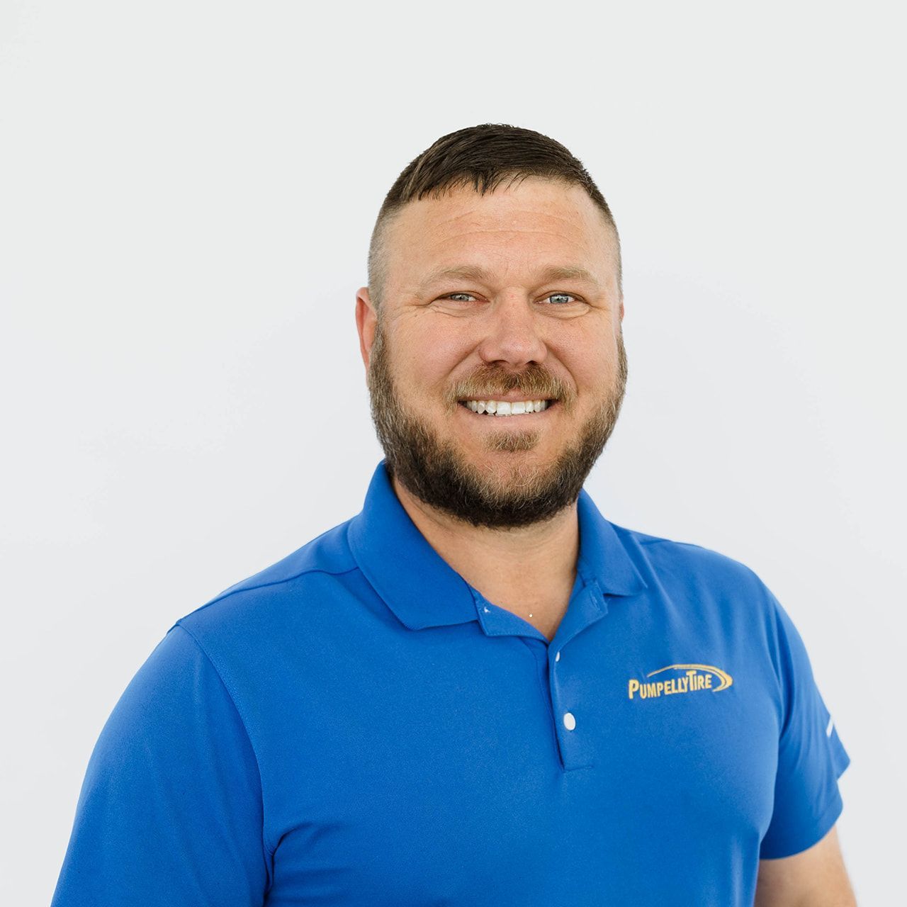 A smiling person with a beard, wearing a blue polo shirt with a gold logo on the chest, posing against a white background.