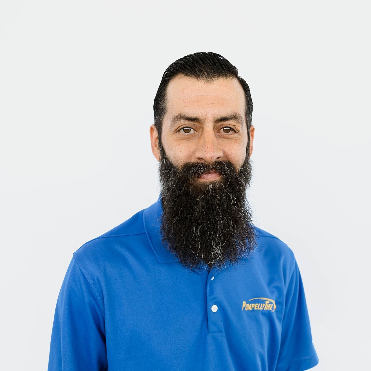 A man with a full beard wearing a blue polo shirt with a logo, standing against a plain white background.