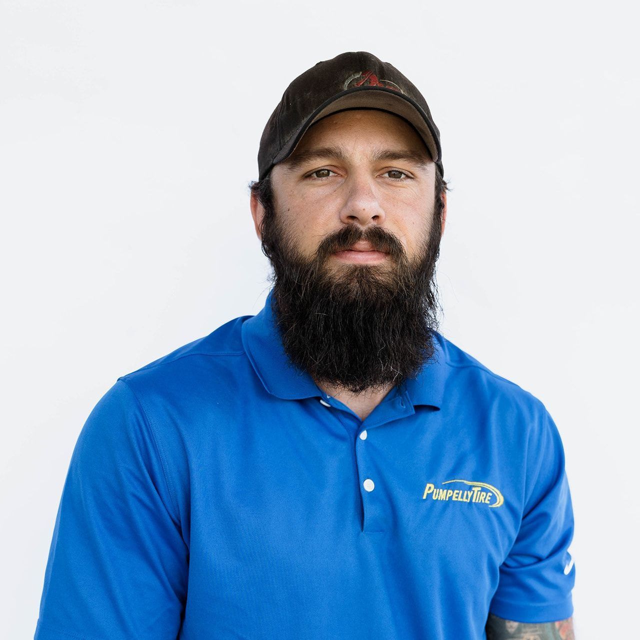 A bearded person wearing a dark cap and blue polo shirt with a logo, standing against a plain white background.