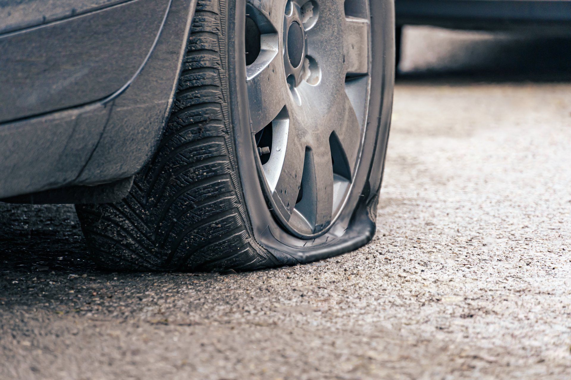 A close-up view of a car's gray wheel and tire that is completely flat, sitting on an asphalt road.
