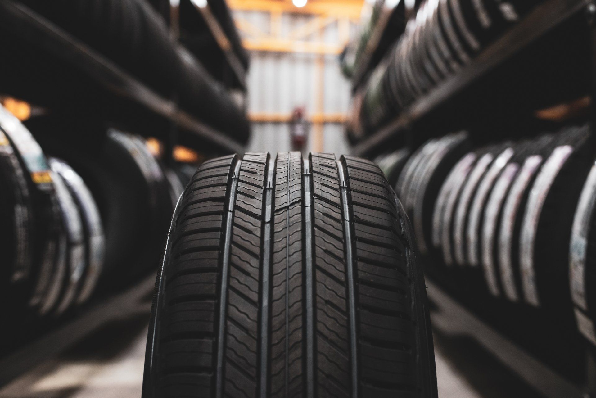 A single car tire in the foreground, centered with a detailed tread, in a dimly lit warehouse filled with stacks of tires.