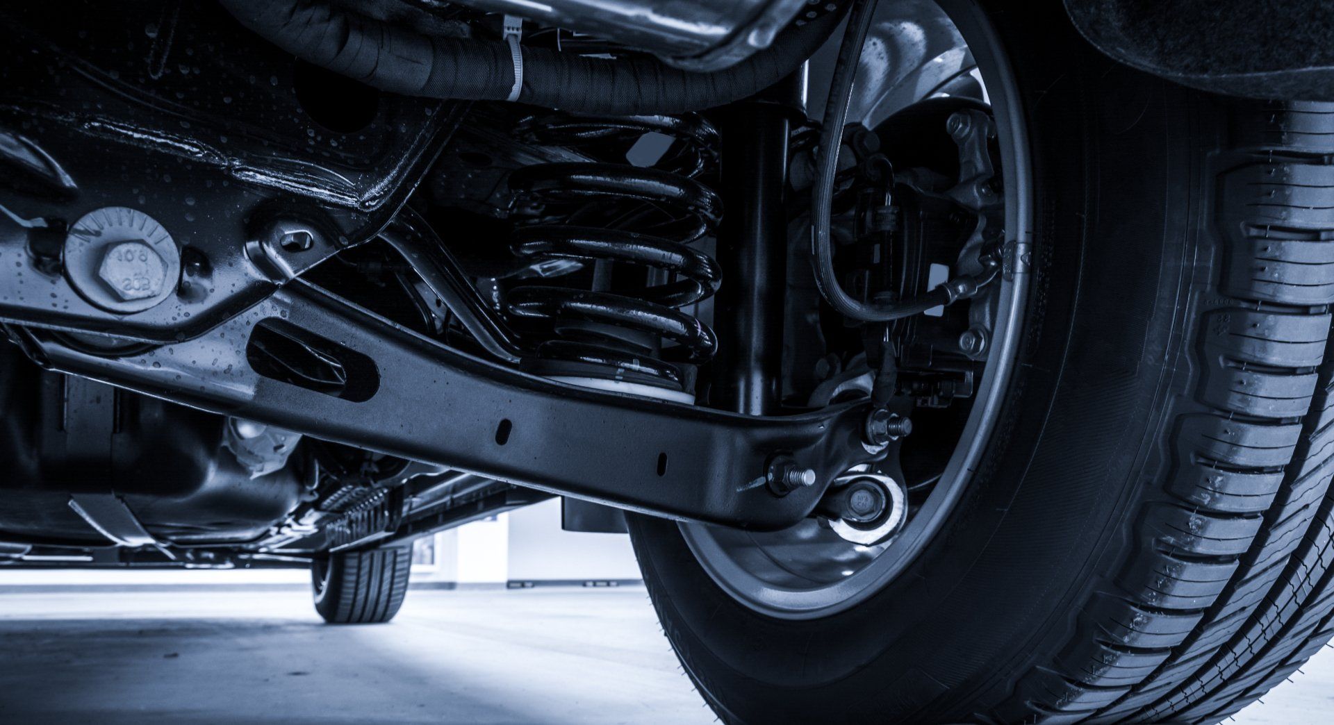 A close-up, low-angle view of a dark automotive vehicle's rear suspension, coil spring, and tire under the chassis.