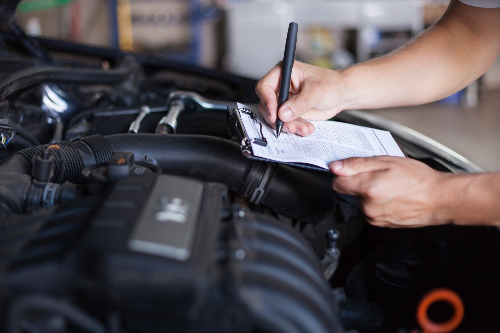 A mechanic writes on a clipboard while inspecting the engine of a car inside a garage.